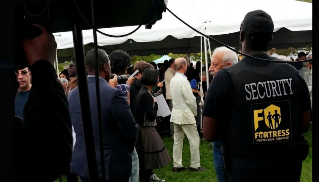 A diverse crowd gathers under a white tent at an outdoor event. A security guard wearing a "Fortress Protection Services" vest stands in the foreground.