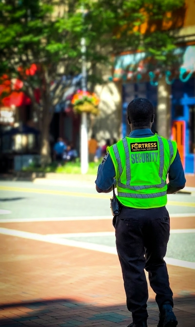 A security guard in a bright green vest walks on a sunny street with trees and colorful storefronts. The scene conveys a sense of safety and vigilance.