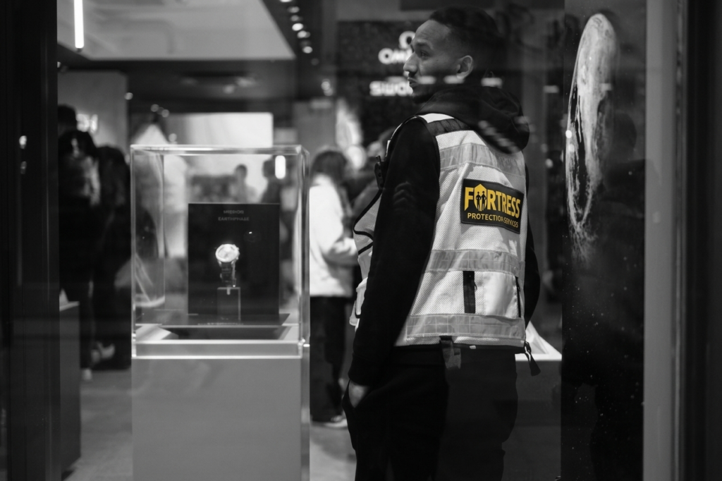 A black-and-white photo shows a security guard in a "Fortress Security" vest, standing near a glass display case holding a wristwatch. A busy store background conveys a sense of alertness.