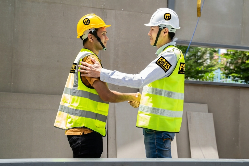 Two construction workers wearing helmets and yellow vests shake hands at a worksite. They appear professional and collaborative, with a positive tone.